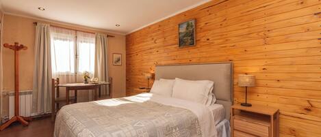 Un dormitorio con pared de madera, una cama, una mesita de noche, una lámpara, una silla, una mesa y una ventana con cortinas.