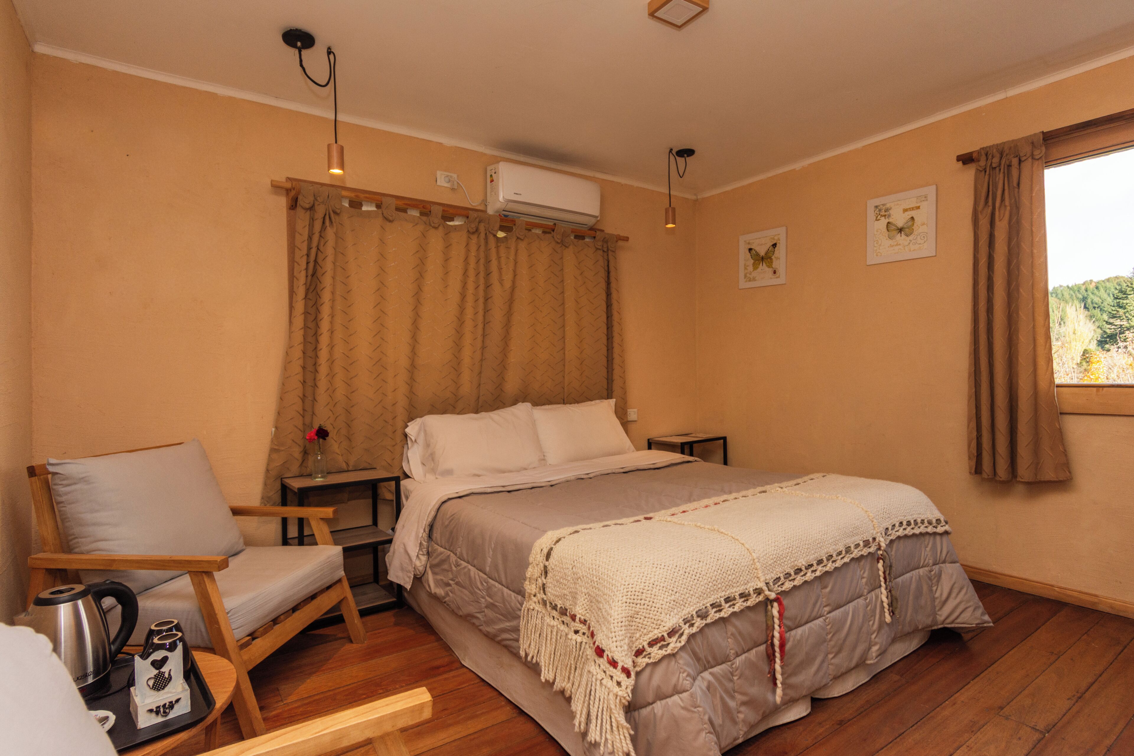 Un dormitorio con cama, una silla, una mesita de noche, un aire acondicionado y una ventana con cortinas.