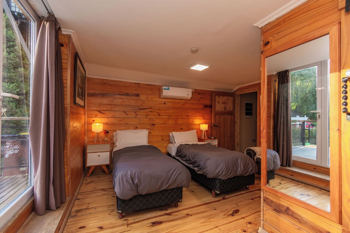 A room with wooden walls, two beds, a window with curtains, a bedside table with a lamp, and an air conditioning unit.