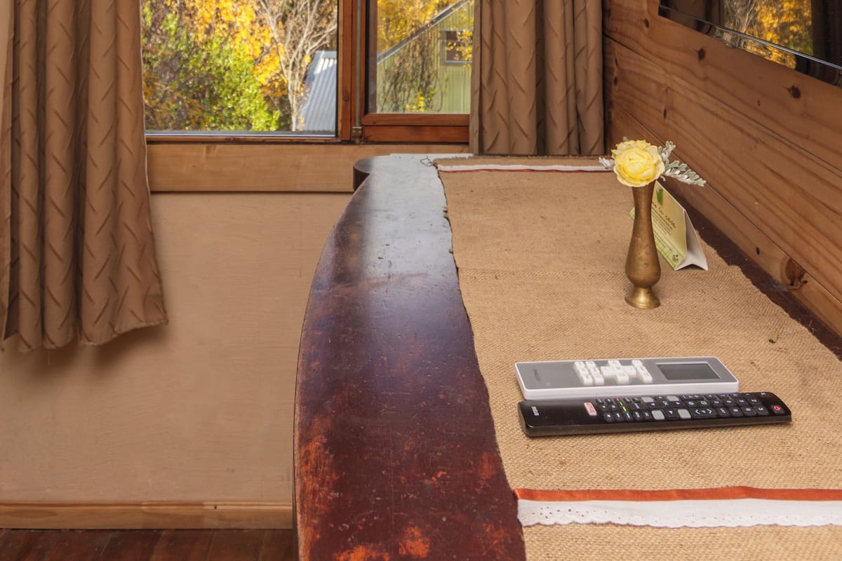 A room with a wooden bench, a remote control, and a vase with a flower on a rug.