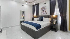 A modern bedroom with a large bed, dark curtains, a wall-mounted artwork, and a bedside table with a lamp. - Benetton Apt - Pure elegance (Lekki, Lagos State, Nigeria)