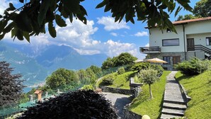 Ein Haus mit einem gepflasterten Weg, der zu einer Holztür führt, umgeben von üppiger Vegetation und Blick auf die Berge.