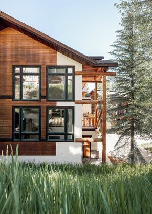 A modern log cabin with large windows, a wooden deck, and a view of a coniferous tree.
