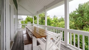 A covered porch with a wooden table and white railings, overlooking a lush green landscape.