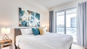 A modern hotel room with a large bed, a city view, and a geometric artwork on the wall.