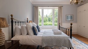 A bedroom with a bed, a nightstand, a lamp, a chandelier, a window with curtains, and a view of greenery outside. - Gorgeous 3BD in Oxfordshire - Stunning Countryside (Steeple Aston)
