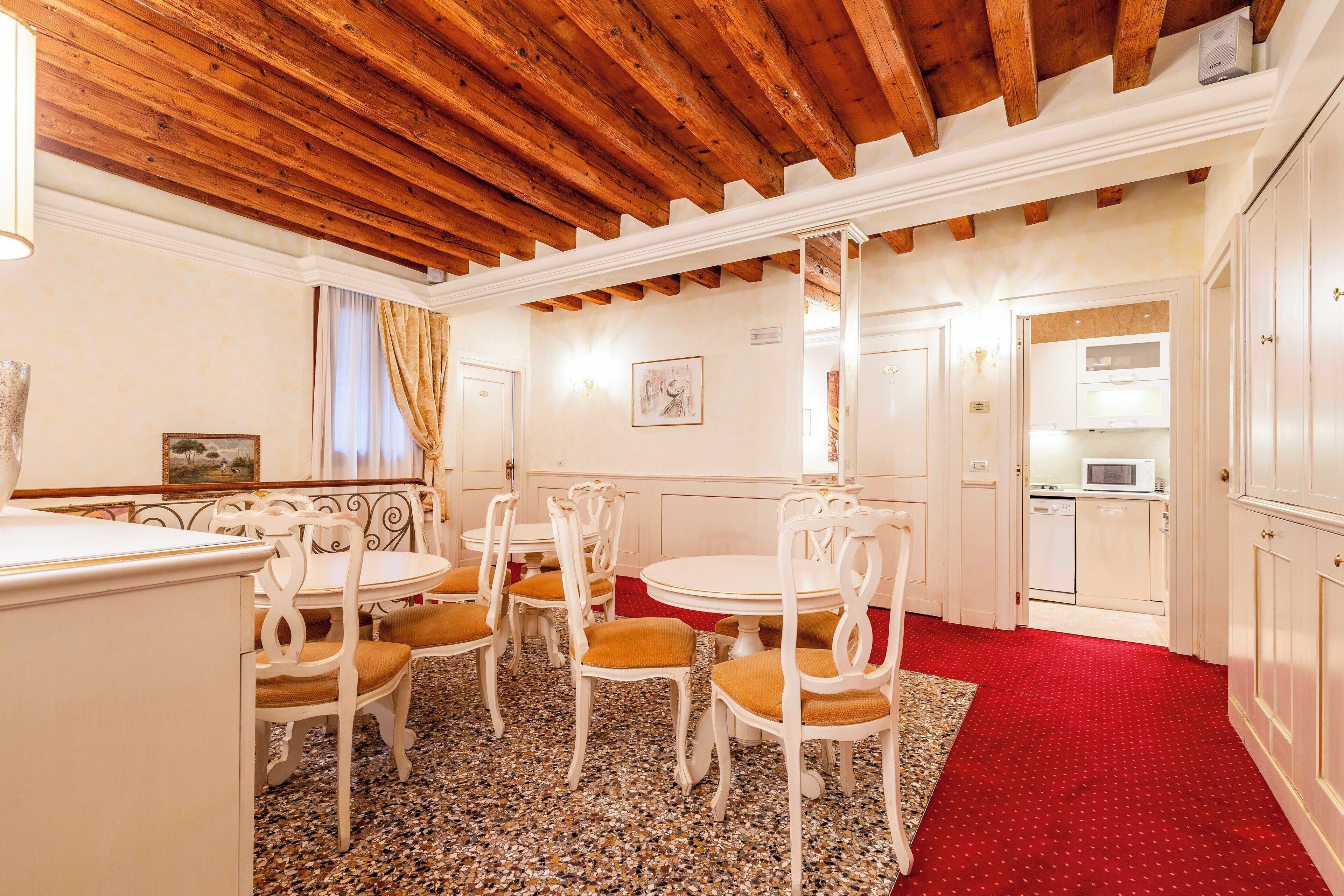 A dining area with white tables and chairs, a red carpet, and a kitchen visible through an open door.