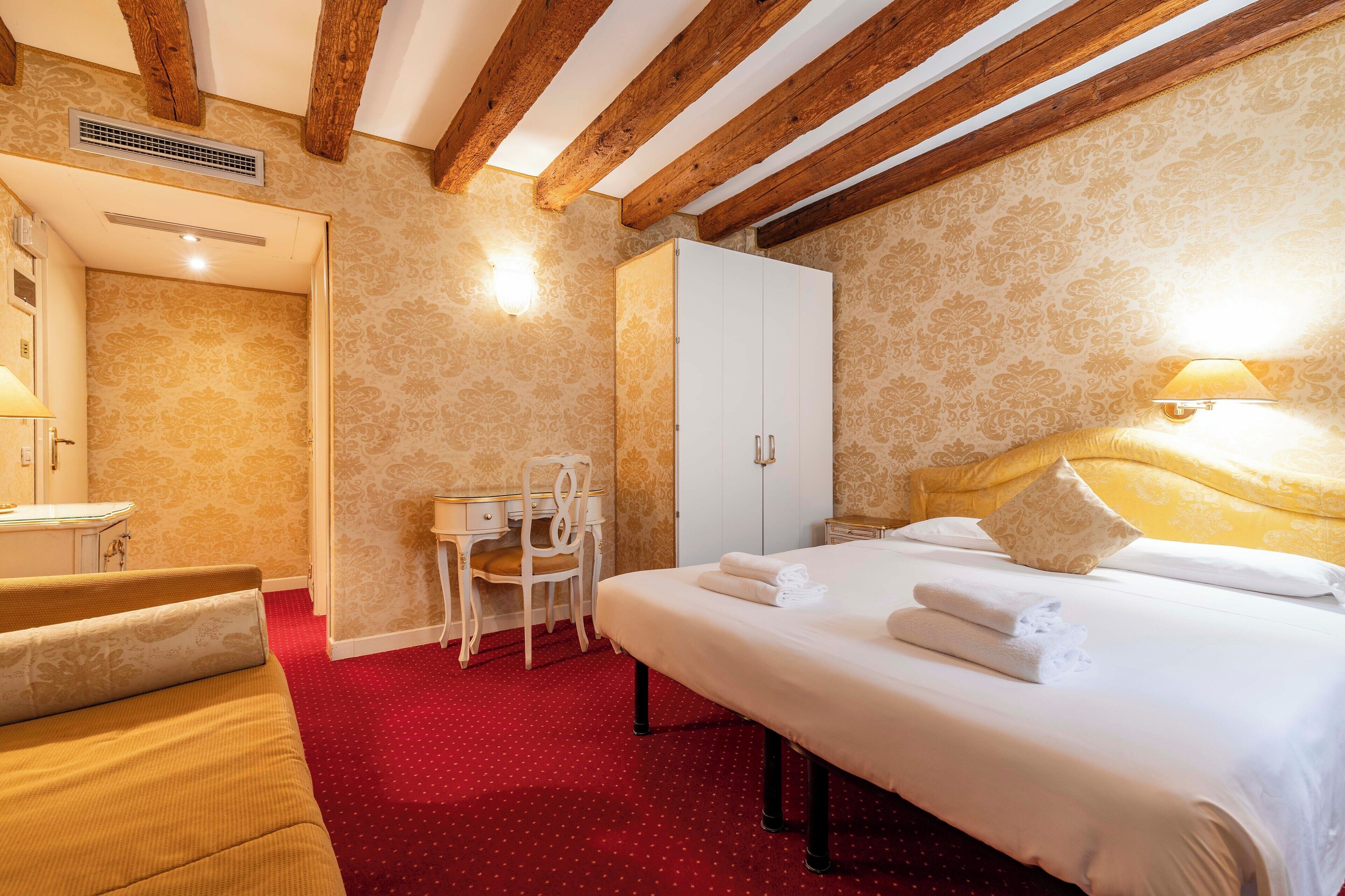 A hotel room with a bed, a desk, a chair, a wardrobe, and a red carpet.