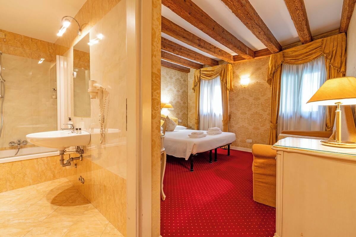 A hotel bathroom with a sink, mirror, and shower. There is a bed with a red carpeted room in the background.