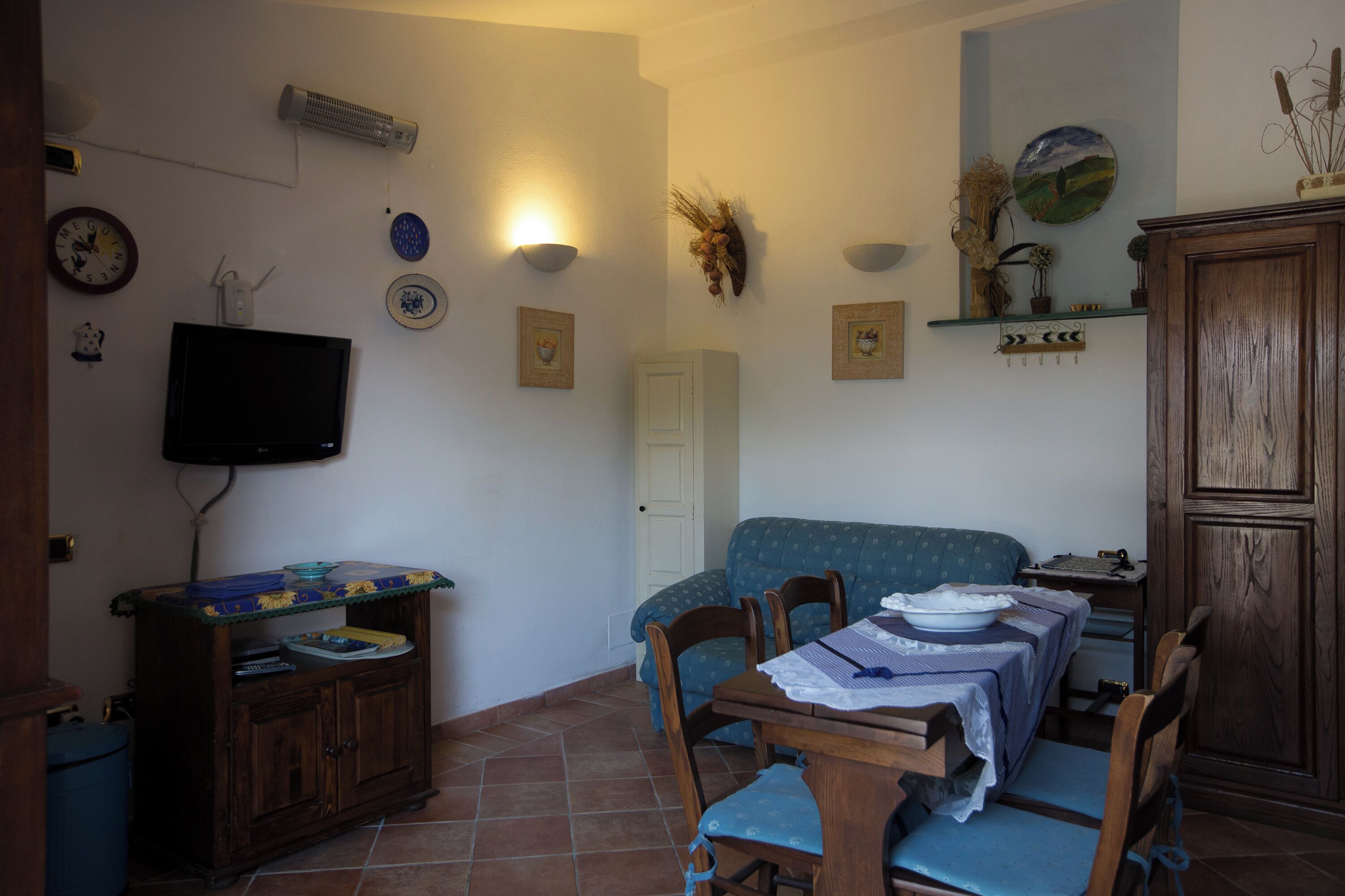 Zona pranzo con tavolo apparecchiato per due persone, televisore a parete e mobile con piano decorativo.