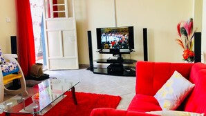 Ein Wohnzimmer mit einem roten Sofa, einem Glastisch, einem Fernseher auf einem Schrank und einer weißen Tür mit rotem Vorhang.