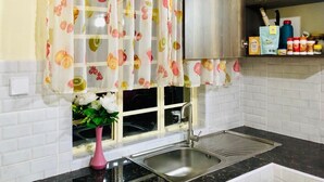 Eine Küche mit einem Edelstahlspülbecken, einem Fenster mit einem fruchtig bedruckten Vorhang, einer rosa Vase mit Blumen und einem Gewürzregal auf der Arbeitsplatte.