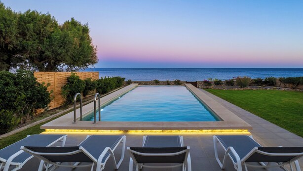 A rectangular swimming pool with a view of the sea, surrounded by a wooden fence and greenery.