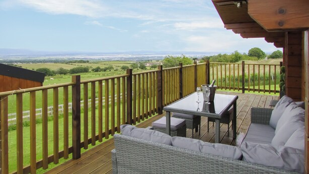 A wooden deck with a table and wicker sofa, overlooking a green landscape. - Stag Lodge - UK46921 (Falkirk)