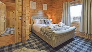 A wooden cabin bedroom with a bed, a nightstand, a window with curtains, and a door leading to another room. - Stag Lodge - UK46921 (Falkirk)
