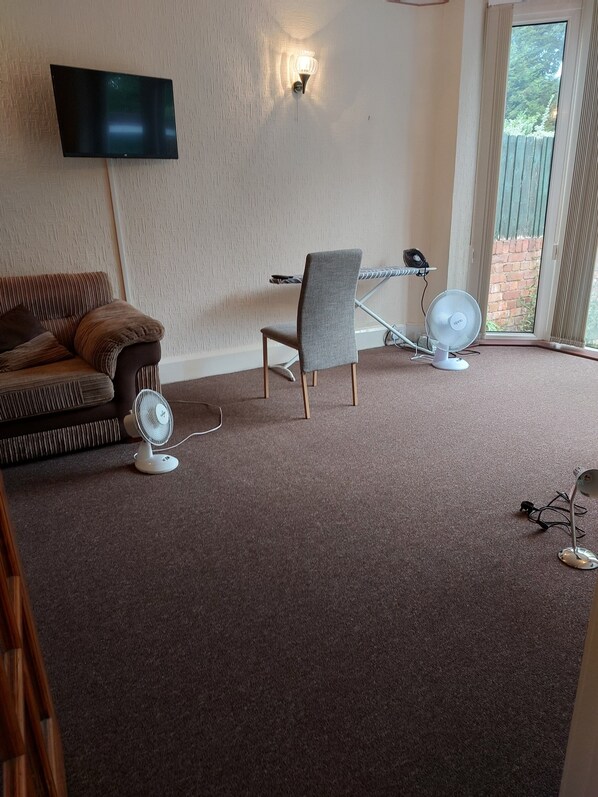 A living room with a flat-screen TV mounted on the wall, a grey upholstered chair, a small table, and two standing fans. - Birmingham Blissful Retreat With 3 Bedrooms (Birmingham)