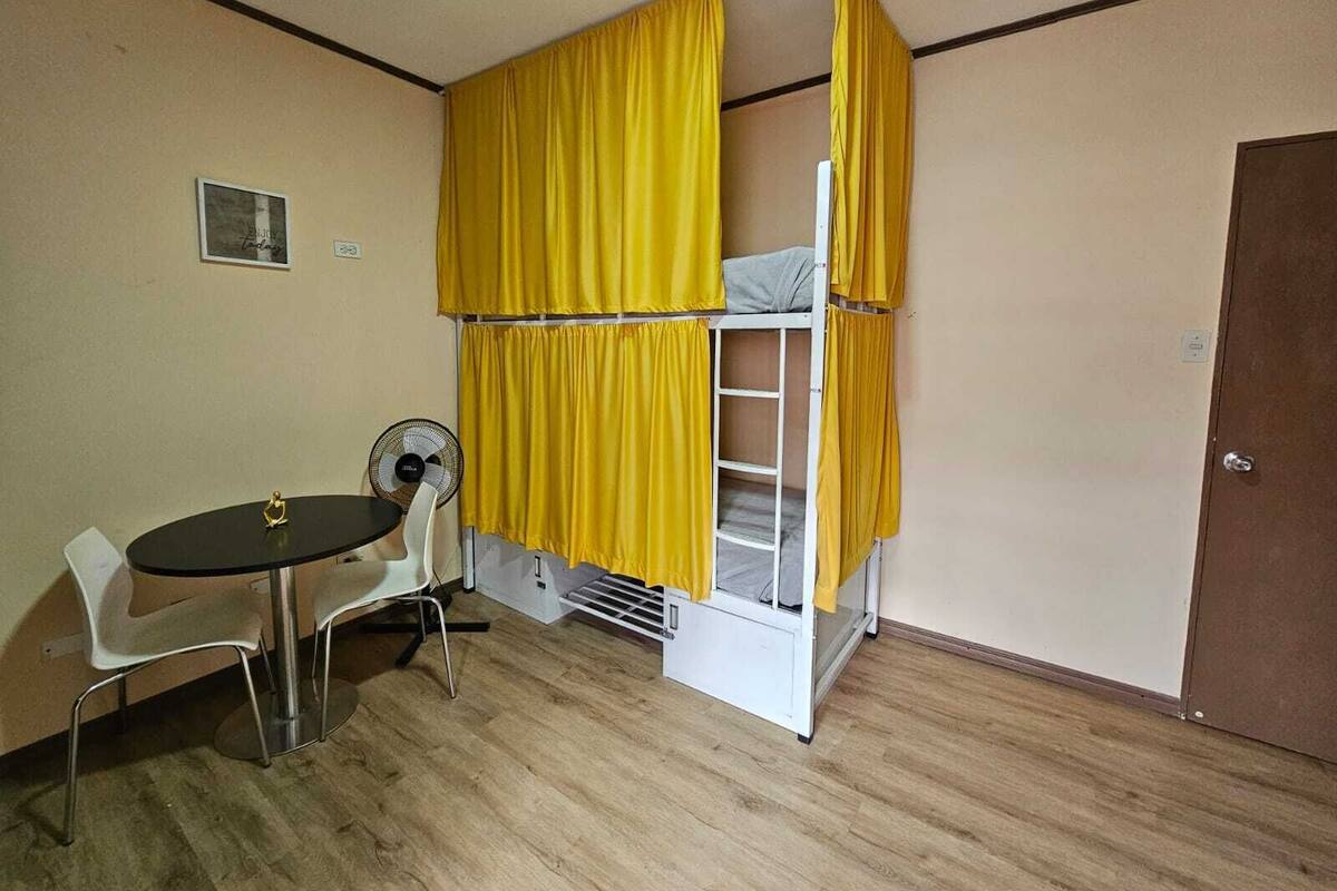A small room with a bunk bed, a table with a chair, a fan, and a door.
