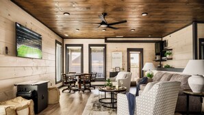 A spacious living room with a wooden ceiling, a ceiling fan, a large TV mounted on the wall, a sofa, a coffee table, and a dining area with chairs and a table.