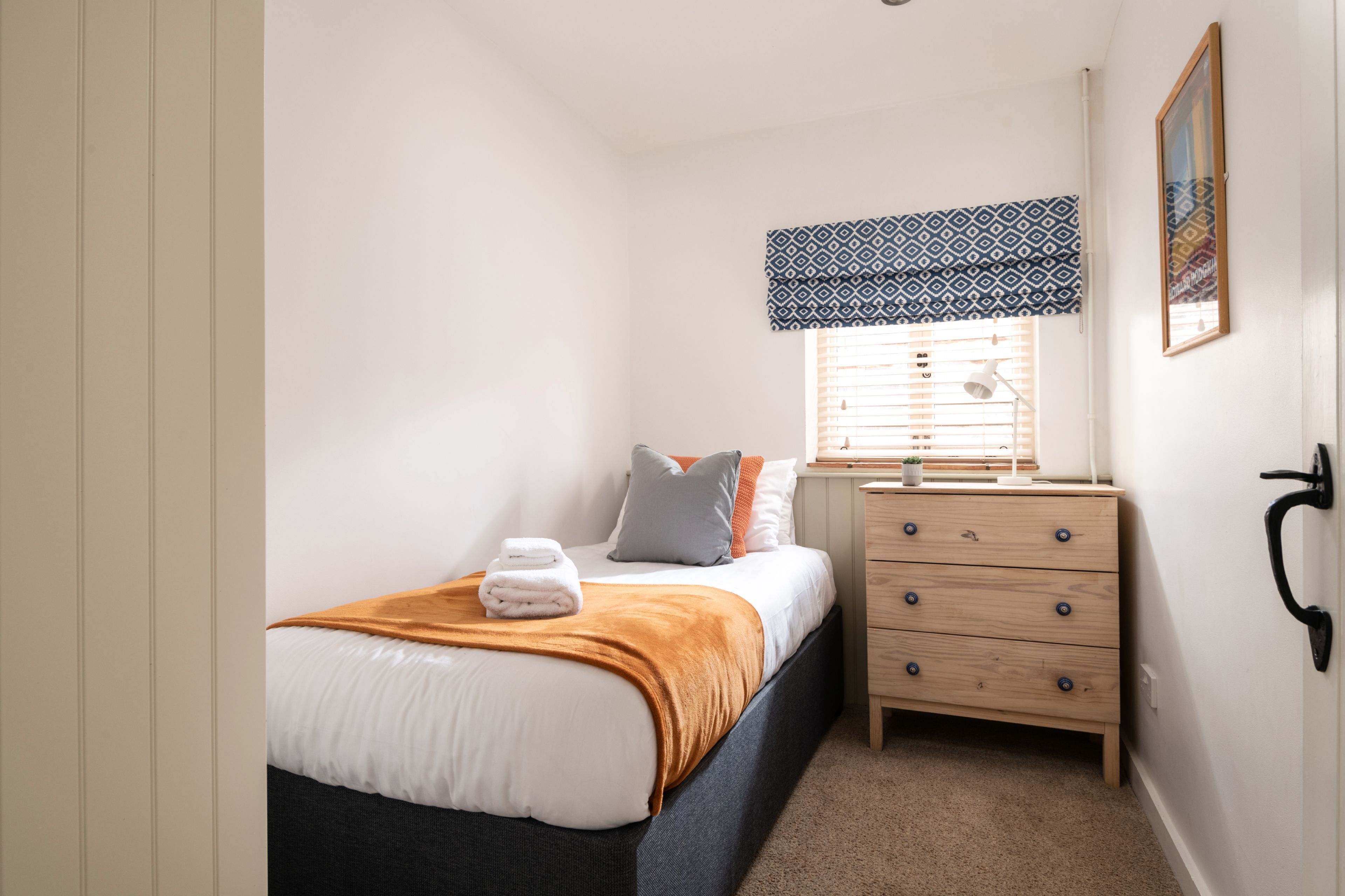 Une chambre à coucher comprenant un lit, une table de chevet, une commode, une fenêtre avec des stores et un tableau encadré au mur.
