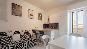 Una cocina moderna con armarios blancos, encimera negra y estufa integrada. Hay una mesa de comedor blanca con cuatro sillas y un sofá blanco y negro con cojines.