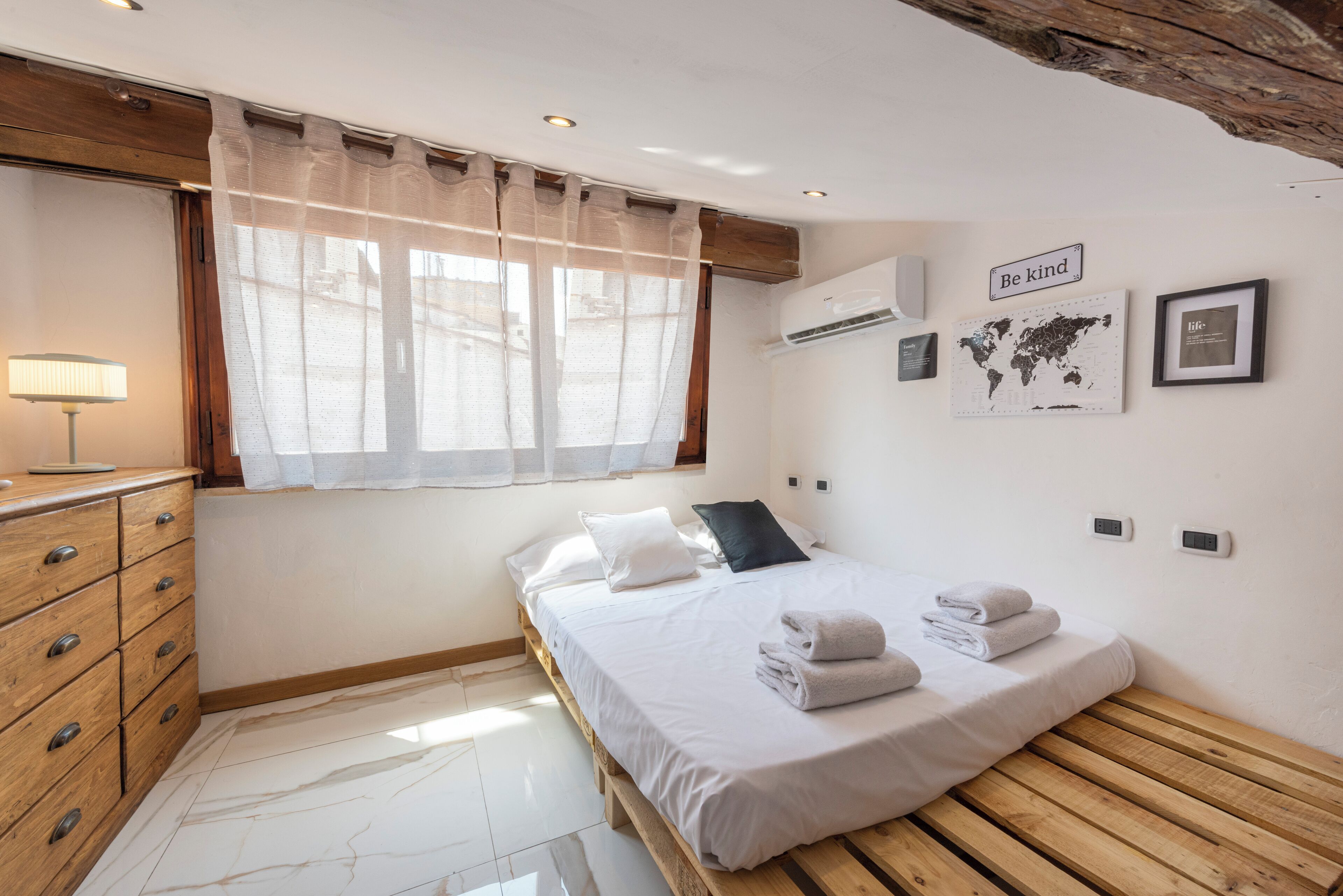 A bedroom with a wooden bed, a bedside table, a lamp, and a window with sheer curtains.