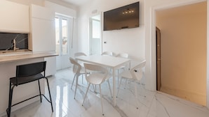 Una cocina moderna con armarios blancos, una encimera de mármol negro y una estufa empotrada. Hay una mesa de comedor blanca con seis sillas y un televisor montado en la pared.