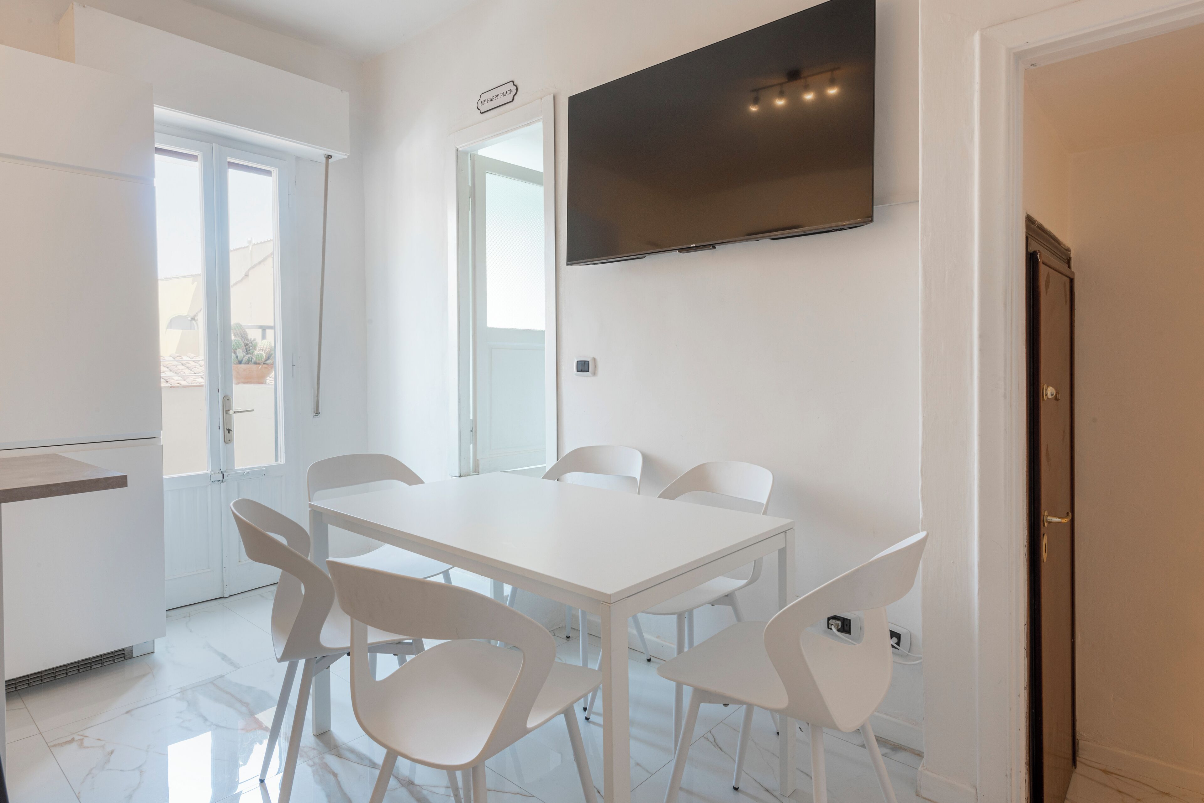 A modern, well-lit room with a white dining table and chairs, a flat-screen TV mounted on the wall, and a door leading to an outdoor area.