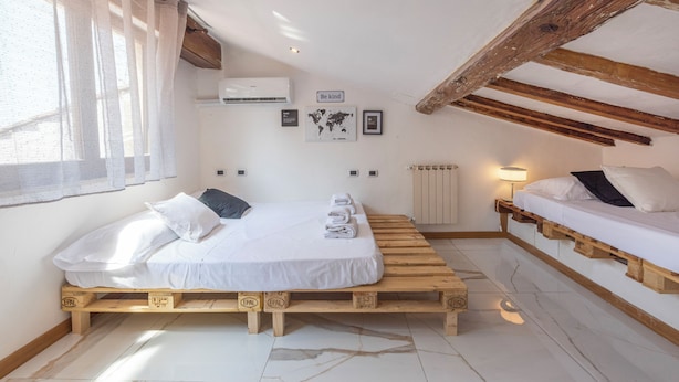 Un dormitorio con cama de madera, ventana con cortinas translúcidas, equipo de aire acondicionado, lámpara de pared y un cuadro con un mapa del mundo.
