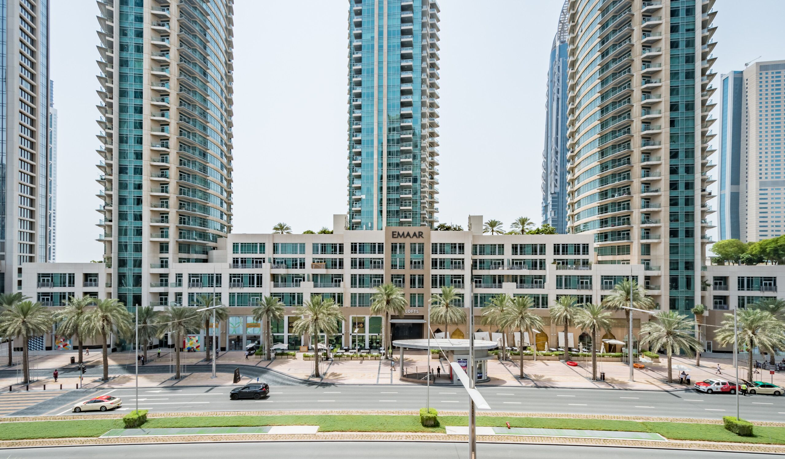Um edifício moderno com vários andares, um sinal EMAAR bem visível e uma praça arborizada com palmeiras.