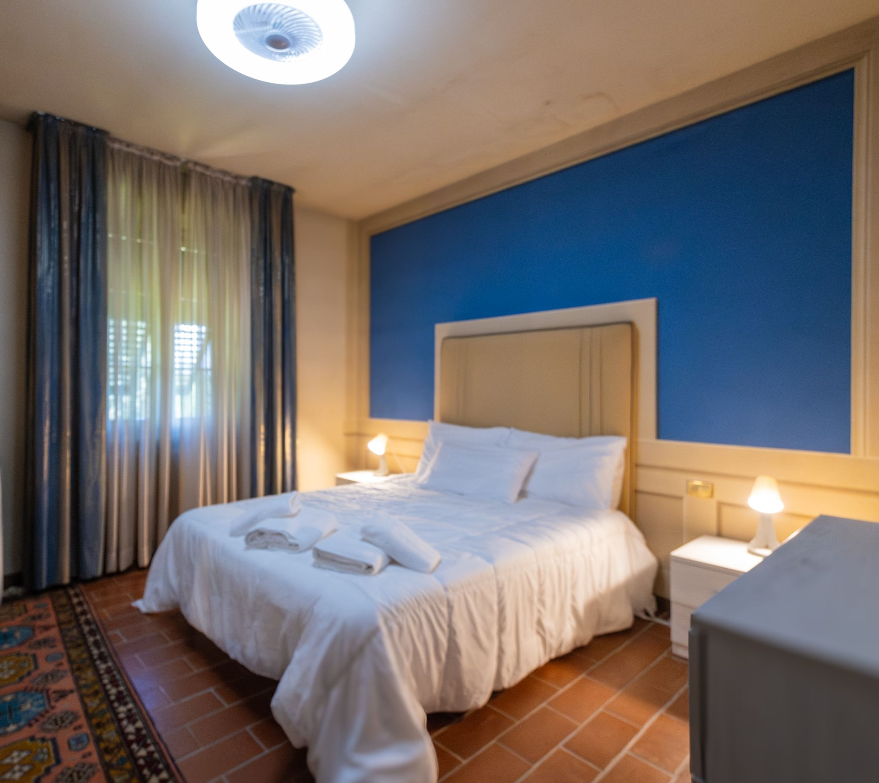 A neatly made bed with white linens and towels, a bedside table with a lamp, and a blue accent wall.