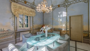 A dining area with a glass table surrounded by patterned chairs, a chandelier, and a window with a decorative valance. - Eighteenth-century Tuscan Villa Tricolle (Fucecchio)