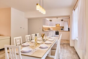 Un comedor moderno con una mesa larga dispuesta para seis personas, sillas blancas y una cocina al fondo.