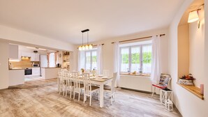 Ein geräumiger Essbereich mit Holztisch und -stühlen, im Hintergrund eine Küche und große Fenster mit Vorhängen.