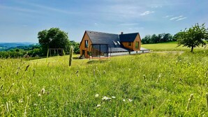 Ein modernes Haus mit geneigtem Dach und Holzfassade, umgeben von einer grünen Wiese und Bäumen.