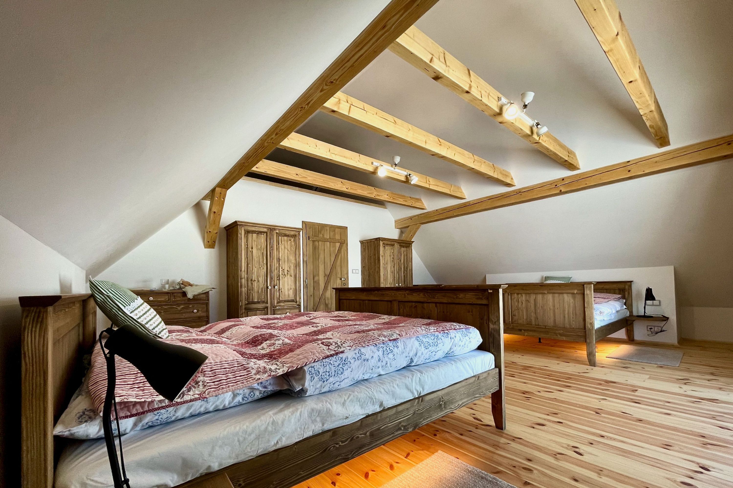 A bedroom with a sloped ceiling, wooden beams, a bed with a patterned bedspread, a nightstand, and a dresser.