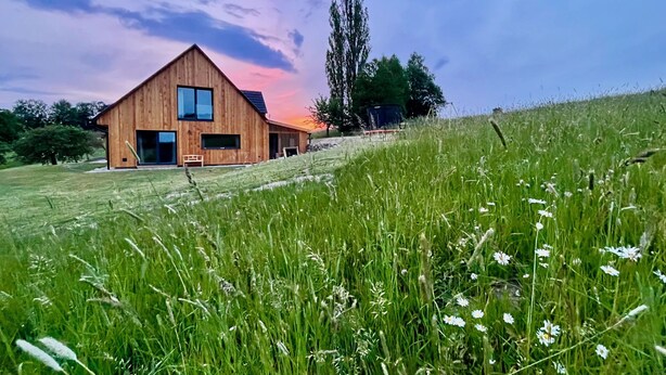 Ein Holzhaus mit einem großen Fenster und einer Tür, umgeben von einem grasbewachsenen Feld mit Wildblumen.