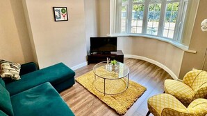 Un salón con un sofá verde oscuro, sillas estampadas de color amarillo, una mesa de centro de vidrio y un televisor sobre un mueble.