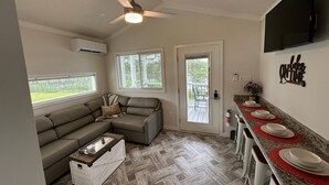 A compact living space with a sofa, a coffee table, a ceiling fan, a kitchen area with a countertop and red placemats, and a door leading to a balcony. - Lake View Cabin, 2 Bedrooms, 13 miles from Jacksonville! (Maysville)
