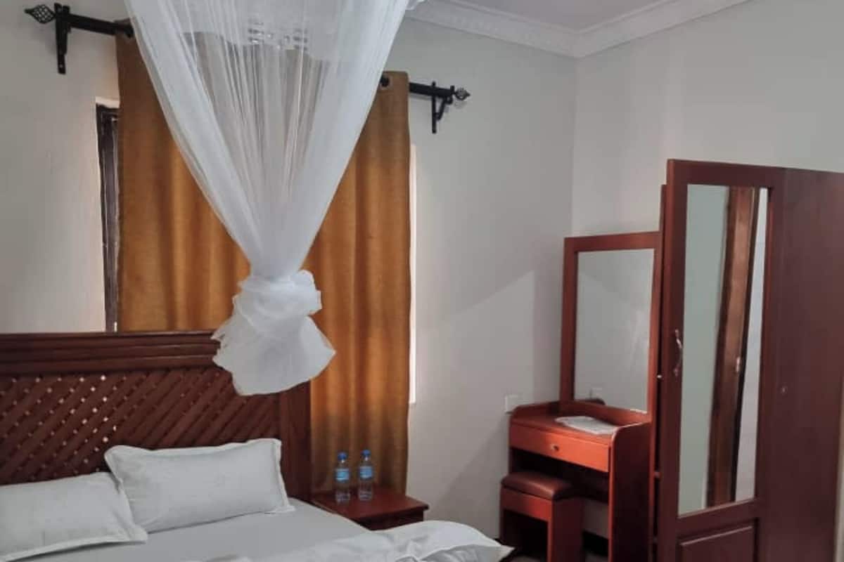 Una camera d'albergo con un letto, un comodino, una cassettiera in legno, uno specchio e un baldacchino sopra il letto.