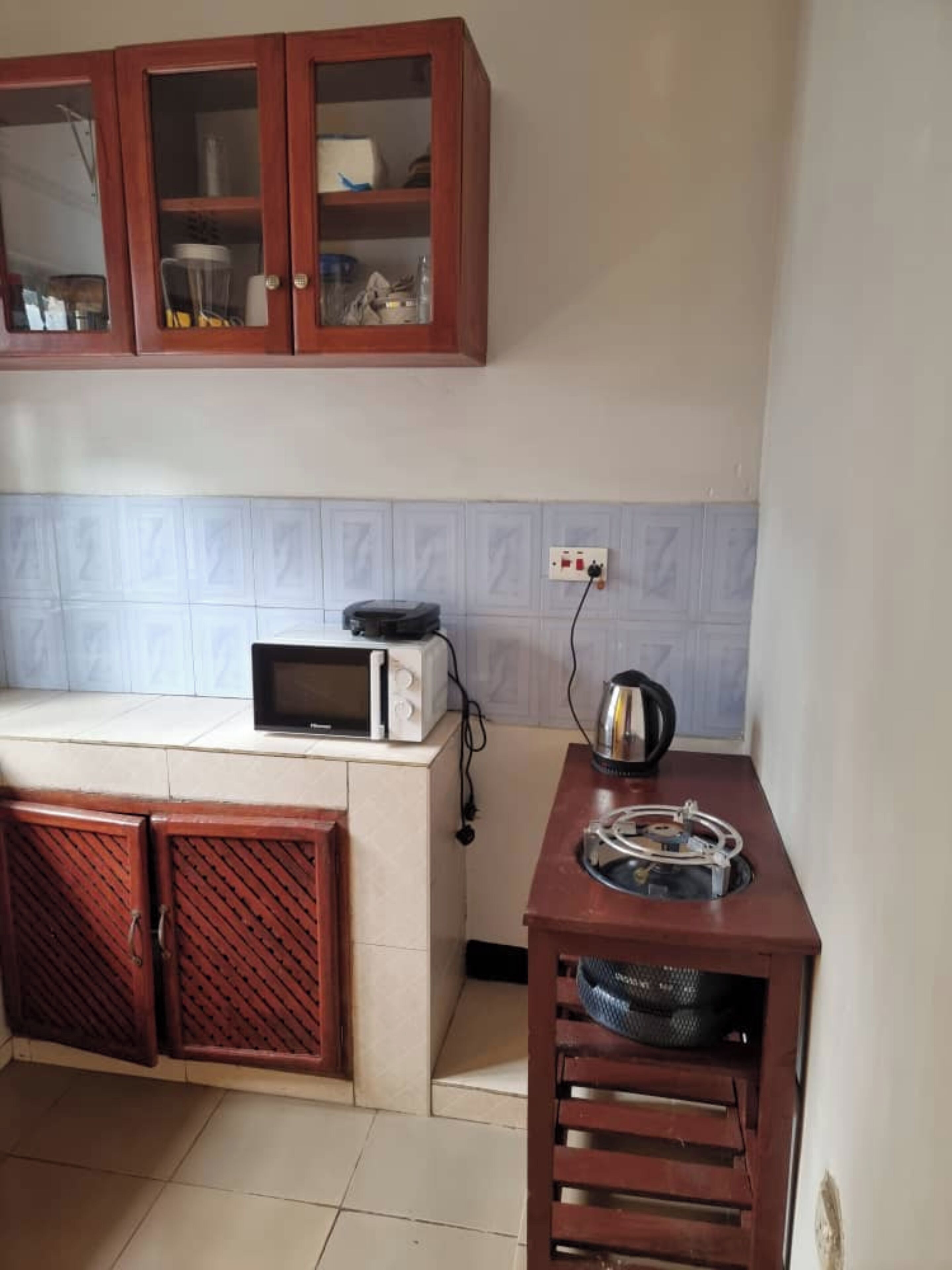 A kitchen with wooden cabinets, a microwave, a kettle, and a small shelf with a basket.