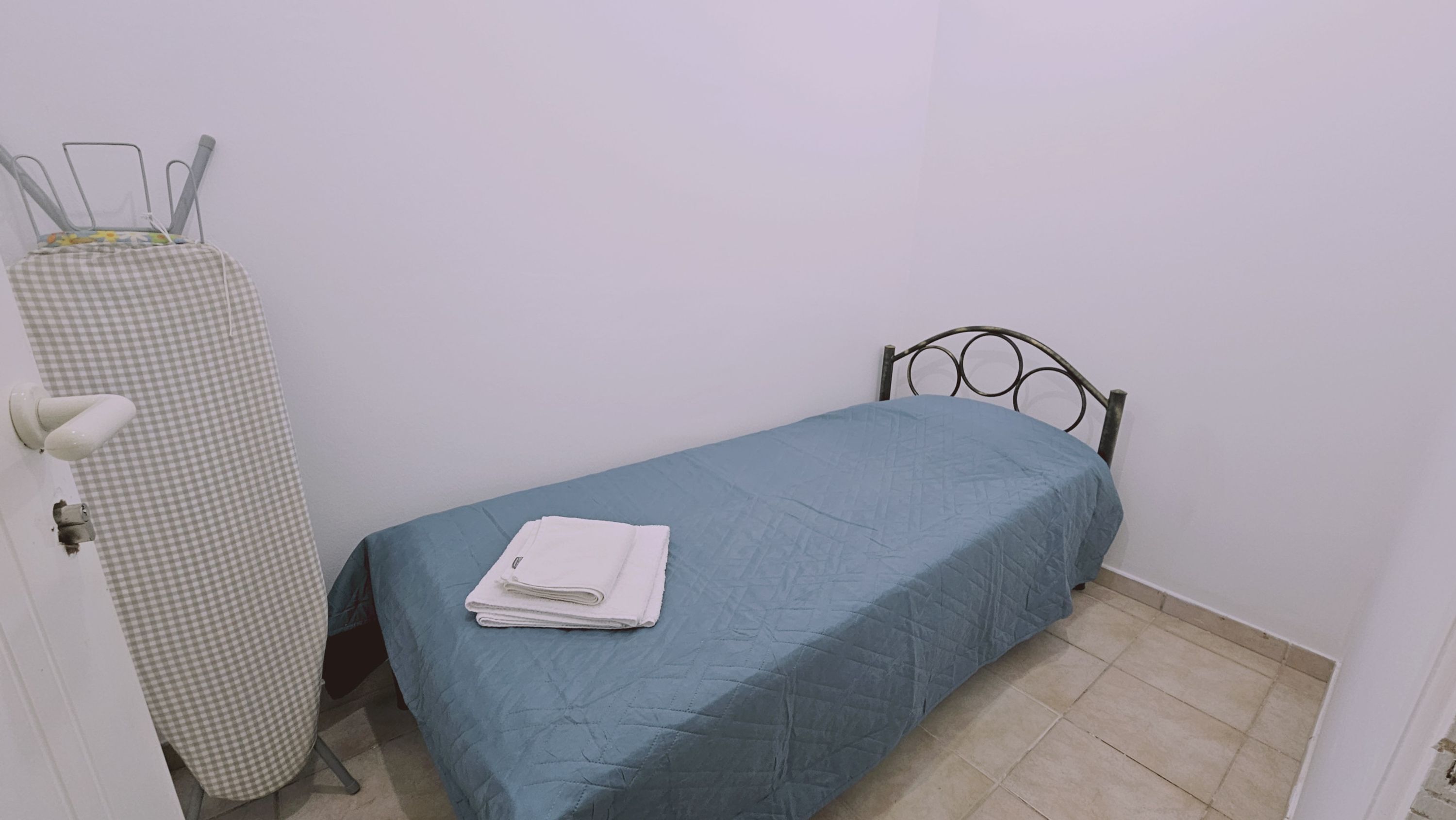 Een kleine eenpersoonskamer met een blauw bedlaken, een metalen bedframe, een geruit strijkplank en een witte handdoek op het bed.