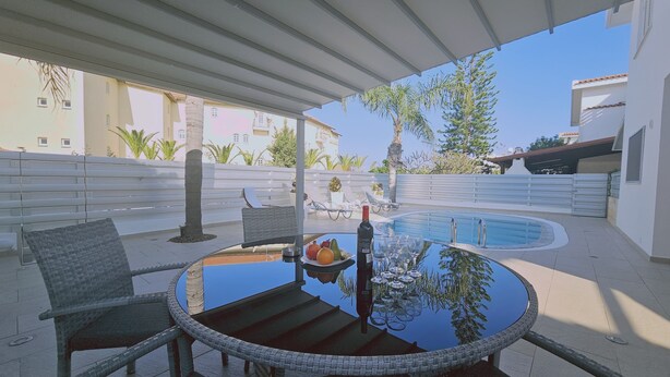 A poolside area with a round table, chairs, and a bottle of wine. - 124 PRO 1 - Stay Villa Prince (Paralimni)