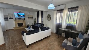 A living room with a white sofa, a small table, a TV, a fireplace, and a window with curtains. - 124 PRO 1 - Stay Villa Prince (Paralimni)