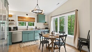 Une cuisine moderne avec des meubles de couleur turquoise, un coin repas doté d’une table ronde et de grandes fenêtres offrant une vue sur la forêt.