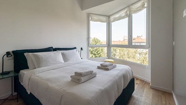 A bedroom with a bed, a nightstand, a lamp, and a window with a view of buildings and trees. - Italianway Comfort - D Aragona 2 (Milan)
