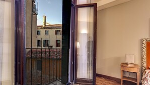 Ein Zimmer mit Holzboden, einem kleinen Holztisch mit Lampe und Blick auf ein Gebäude mit Ziegeldach durch eine offene Tür.