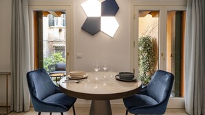 Un coin repas avec une table ronde, deux chaises bleues, un luminaire hexagonal et une vue sur un bâtiment par une porte ouverte.
