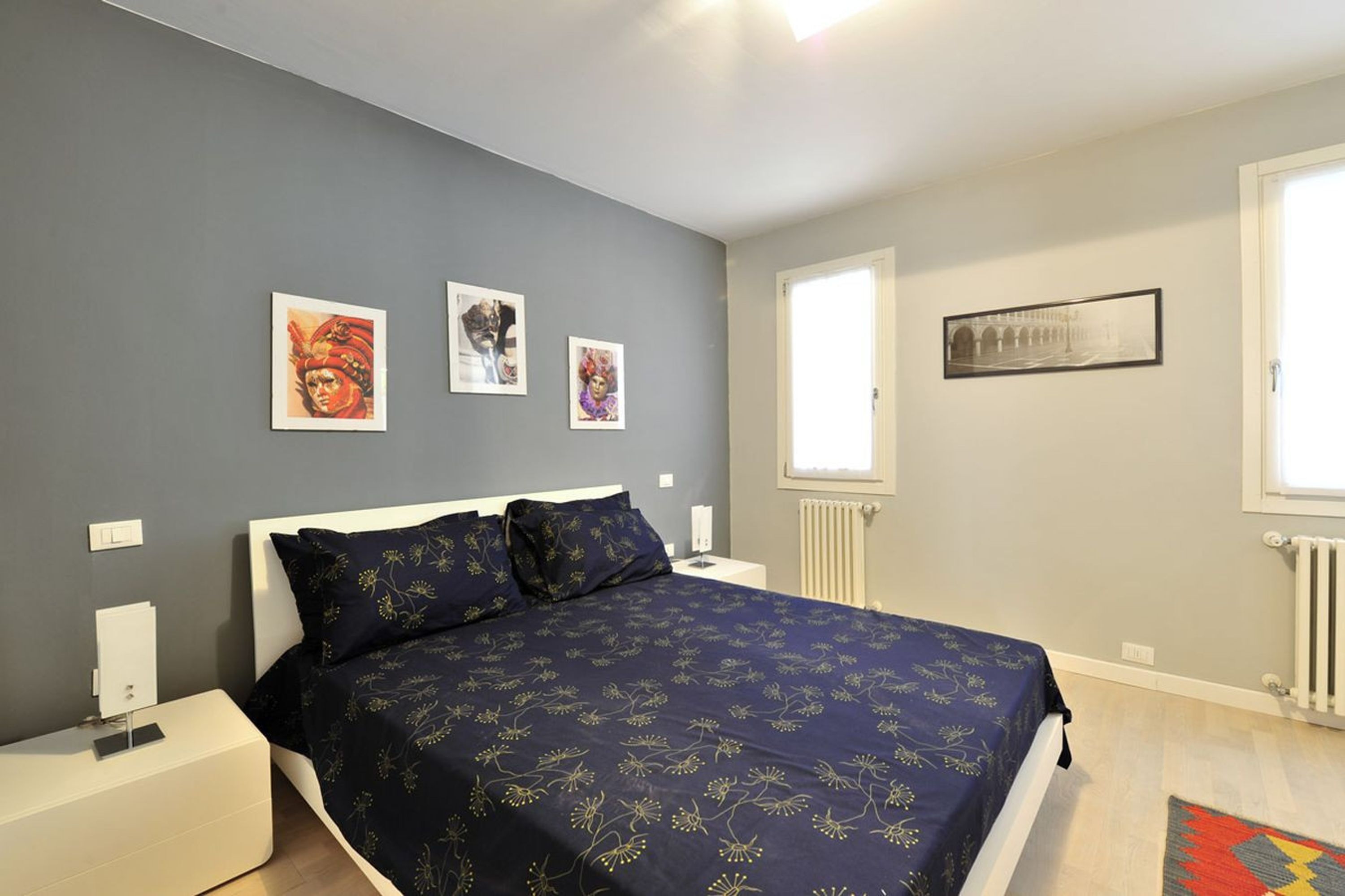 A bedroom with a bed, bedside tables, artwork on the wall, a window, and a radiator.