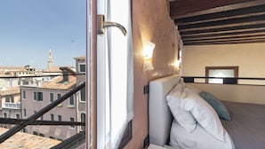 A hotel room with a bed, a window with a view of buildings, and a wooden ceiling. - Doge Palace 8 - Locz (Venice)