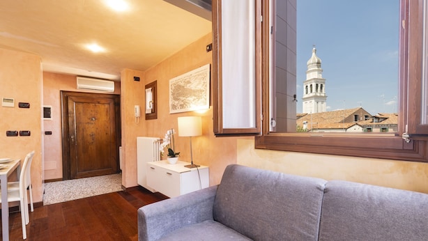 A cozy room with a grey sofa, a wooden door, a white desk with a chair, and a view of a white building with a tower. - Doge Palace 8 - Locz (Venice)